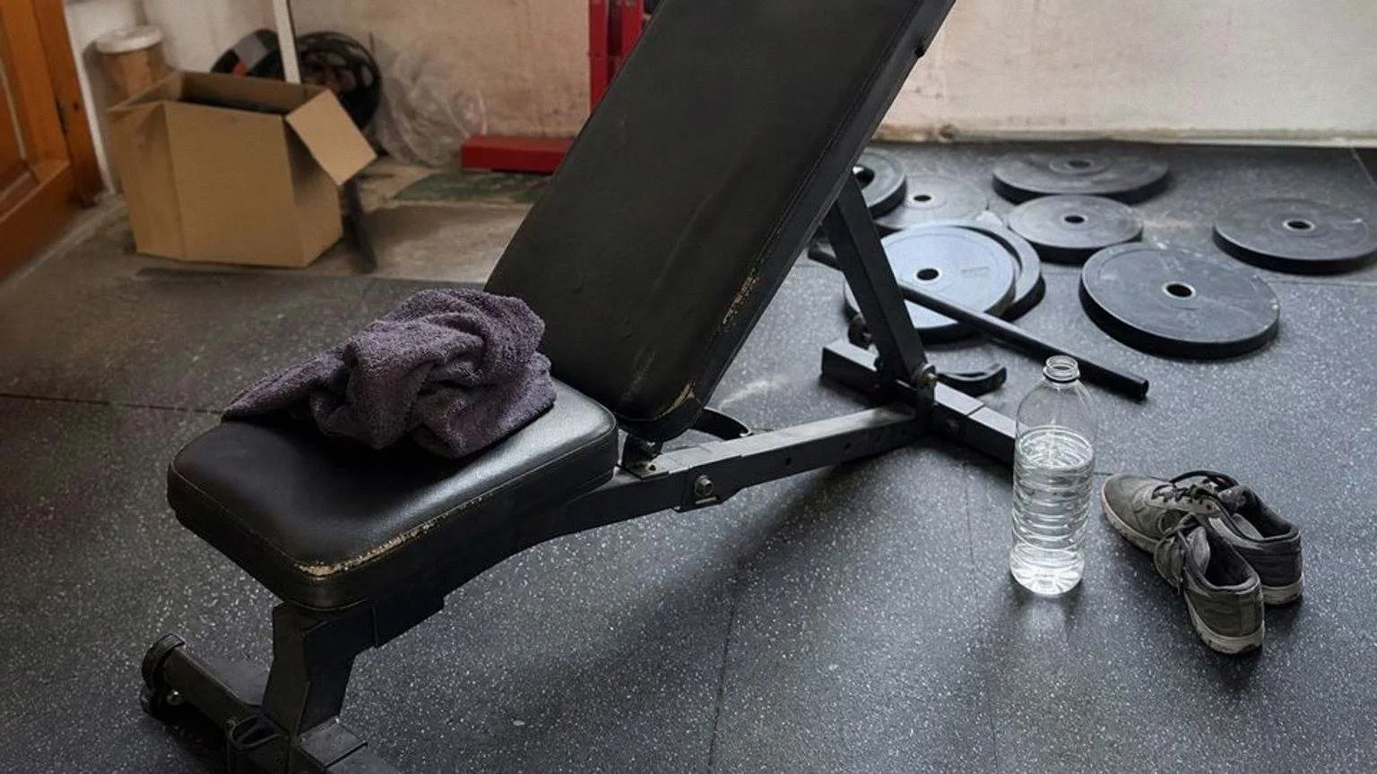 Banco de musculación ajustable con el respaldo inclinado en el rincón de un garaje, con discos de hierro apilados en el suelo de goma y una toalla sobre el asiento.