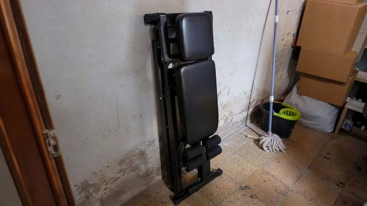 Banco de musculación plegado en vertical y apoyado contra la pared de un trastero, junto a una fregona y cajas apiladas.