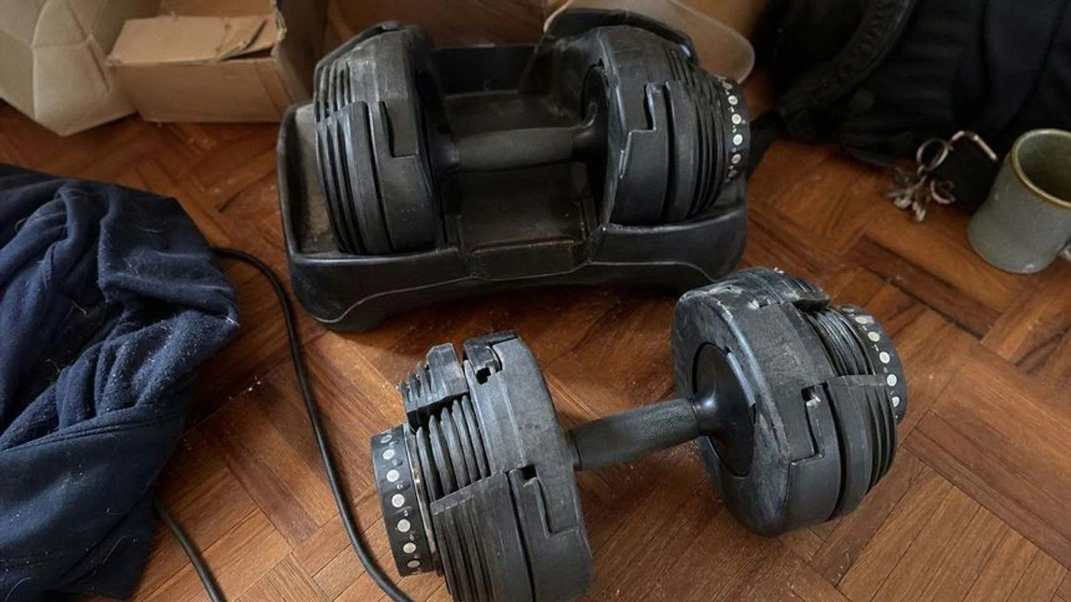 Par de mancuernas ajustables de dial en el suelo de parquet de un salón, una sobre su base y otra fuera, junto a una sudadera arrugada.