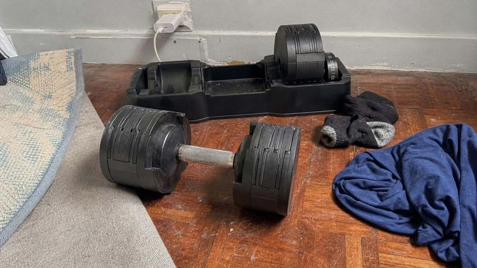 Mancuerna ajustable de dial fuera de su bandeja en el parquet rayado de un dormitorio, con la bandeja y la segunda mancuerna detrás, junto a unos calcetines y una camiseta arrugada.