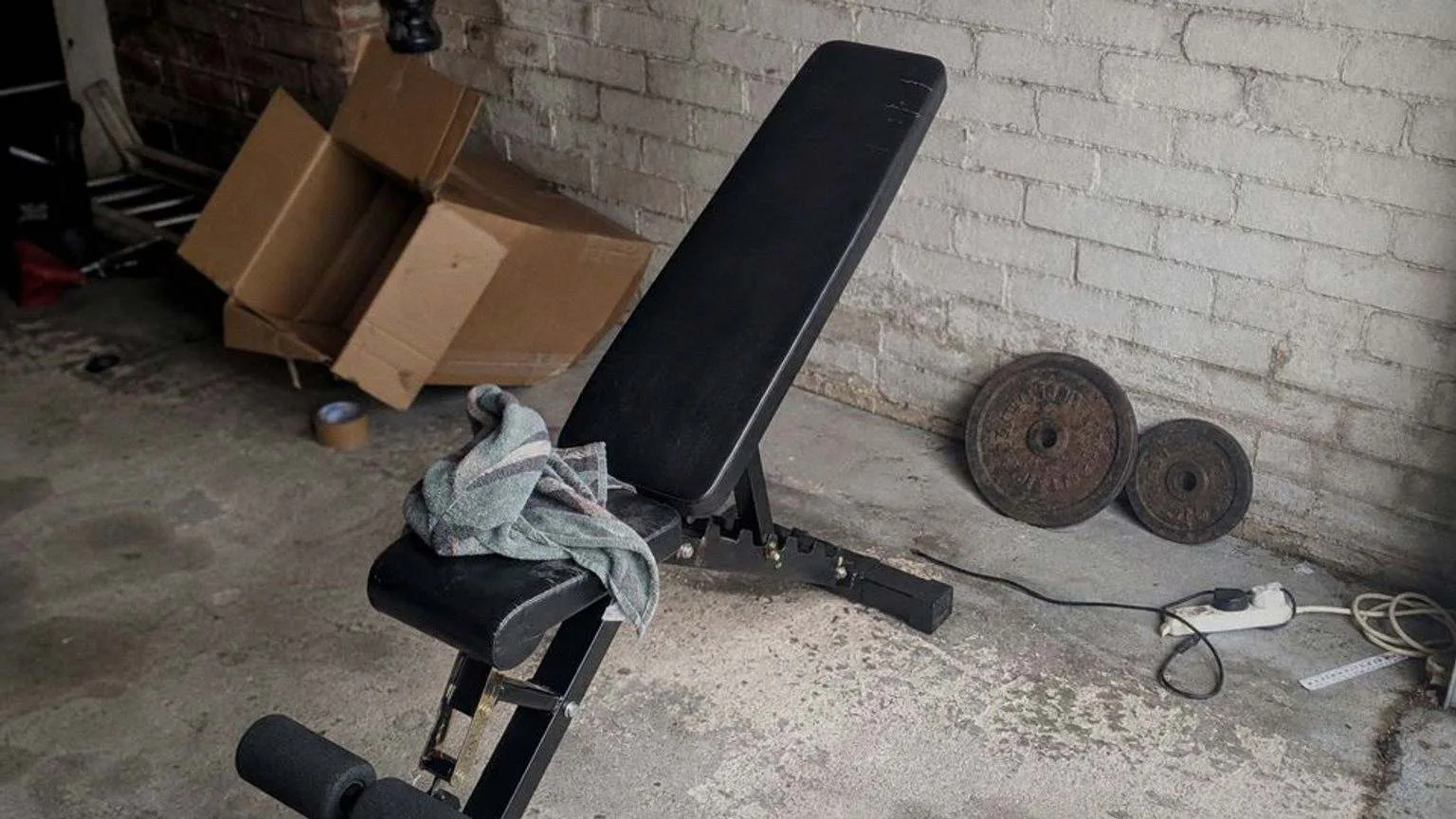 Banco de musculación ajustable montado en el rincón de un garaje con suelo de cemento manchado, una toalla arrugada encima del respaldo y dos discos de hierro apoyados de canto contra la pared.