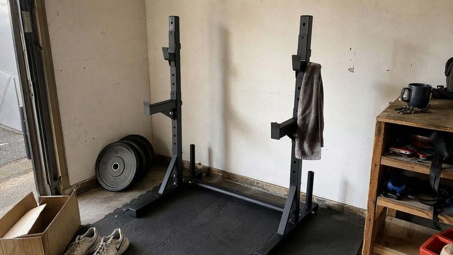 Soporte de sentadillas negro con las dos columnas unidas por un tubo a ras de suelo, montado en la esquina de un garaje sobre esterilla de goma, con discos apoyados en la pared, una toalla colgada de uno de los postes y un banco de trabajo al lado.